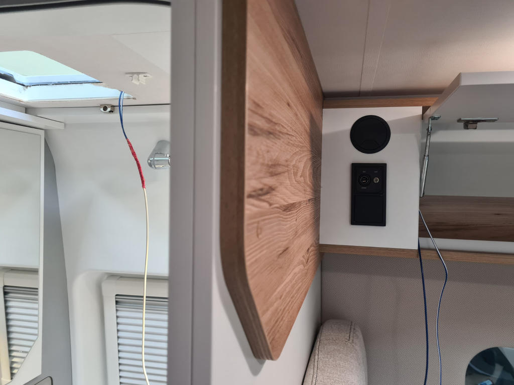 Innenraum mit Holzpaneel, rundem schwarzem Bauteil, schwarzem rechteckigem Bedienelement, sichtbaren blauen, roten und gelben Kabeln, Dachfenster, zwei Fenster mit Jalousien und Polstersitz