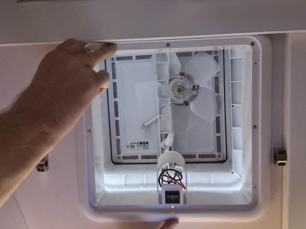 Zwei H&auml;nde halten einen abgenommenen wei&szlig;en L&uuml;ftungsrahmen; darunter sichtbar ein Vierblatt-Ventilator und eine freiliegende Lampe mit roten Dr&auml;hten