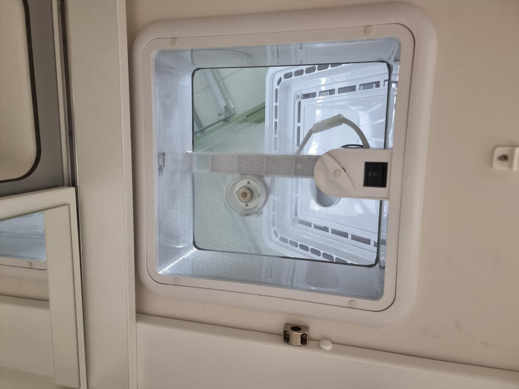 Rechteckige wei&szlig;e Decken&ouml;ffnung mit quadratischem L&uuml;ftungsrahmen, sichtbarem Gitter, zentraler Drehachse und Metallarm mit rundem Knauf; kleines schwarzes Bedienelement, links Schrankkante, unten Verriegelung