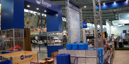 Gro&szlig;er blauer Stand mit Schriftzug Berger, gestapelte Holzkisten und Kartons, blau verpackte Waren und weitere St&auml;nde in einer gro&szlig;en Halle