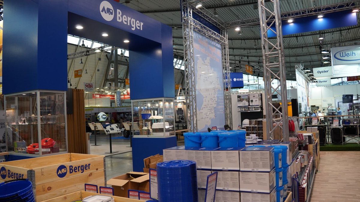 Gro&szlig;er blauer Stand mit Schriftzug Berger, gestapelte Holzkisten und Kartons, blau verpackte Waren und weitere St&auml;nde in einer gro&szlig;en Halle