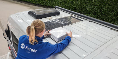 Person with blond ponytail wearing a blue jacket with 'Berger' logo leaning over the roof of a white van, hands on a white rectangular roof vent; two other roof openings and a black rooftop unit visible; hedge along the road