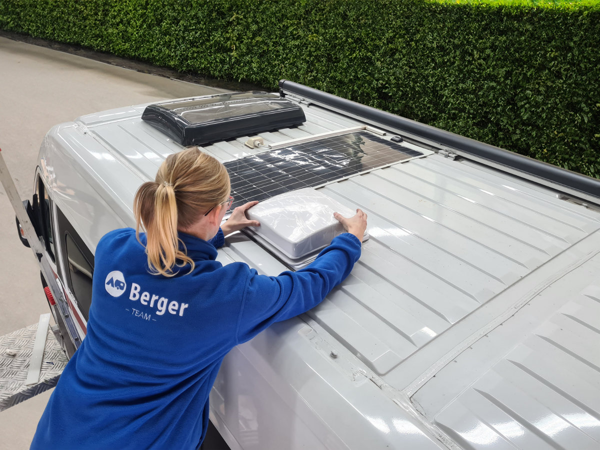 Person mit blondem Pferdeschwanz in blauer Jacke mit 'Berger'-Logo, &uuml;ber das Dach eines wei&szlig;en Vans gebeugt, H&auml;nde auf einem wei&szlig;en rechteckigen Dachl&uuml;fter; zwei weitere Dach&ouml;ffnungen und ein schwarzes Aufsatzger&auml;t sichtbar; Hecke am Stra&szlig;enrand