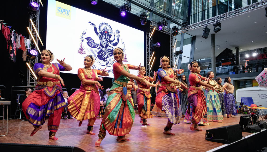 Mehrere Personen in bunten Tanzkost&uuml;men auf einer B&uuml;hne, im Hintergrund ein gro&szlig;er Bildschirm mit dekorativem Motiv und B&uuml;hnenbeleuchtung