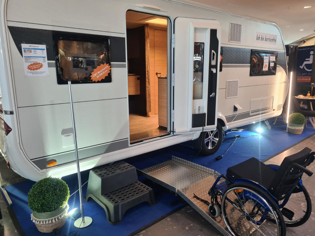 Wei&szlig;er Wohnwagen in Ausstellung, ge&ouml;ffnete T&uuml;r, Aluminiumrampe, Rollstuhl an der Rampe, Plastiktreppen, Topfpflanzen, blauer Teppich, Innenraum mit Holzschr&auml;nken sichtbar, Aufkleber an Fenstern, Beleuchtung