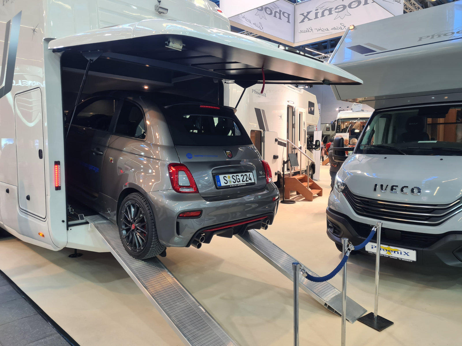 Graues Kleinwagen steht auf Rampen im Heckgaragenbereich eines Wohnmobils; weiteres Wohnmobil, Absperrungen und Personen im Hintergrund