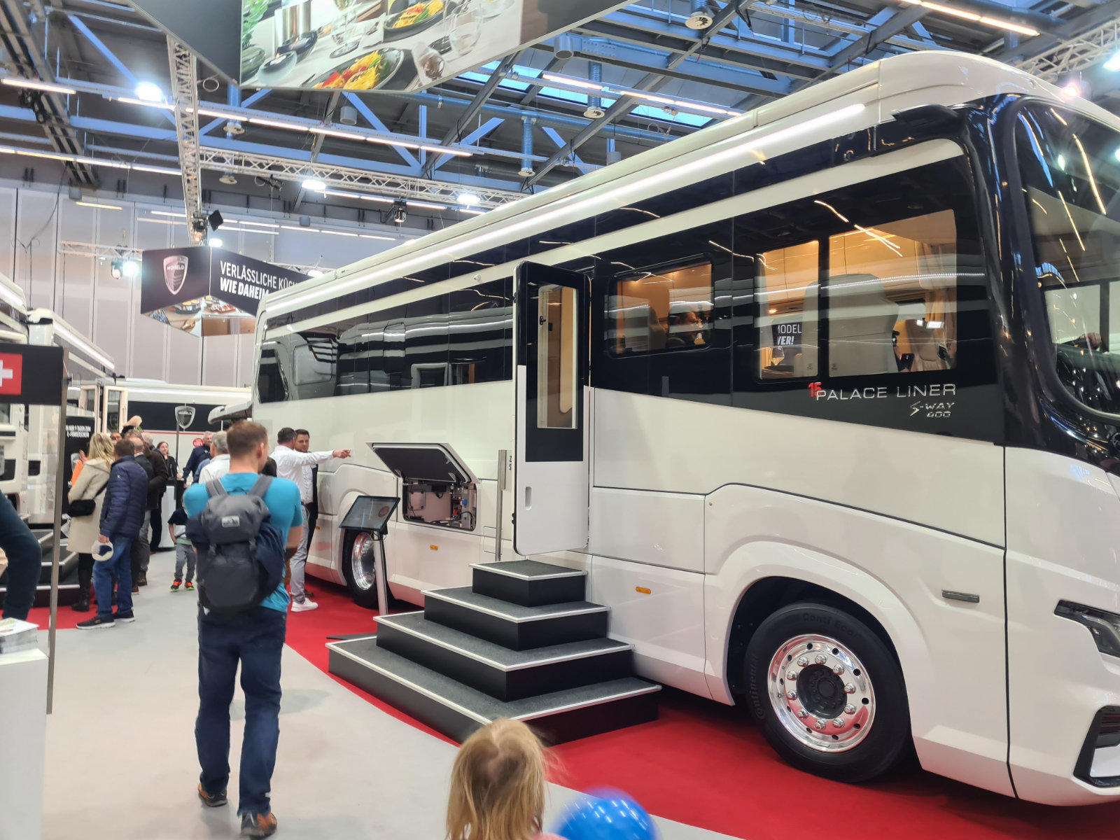 Gro&szlig;es wei&szlig;es Wohnmobil in einer Ausstellungshalle, ge&ouml;ffnete Seitenklappe, mobile Treppe zur T&uuml;r, mehrere Personen davor, sichtbare Hallendecke mit Beleuchtung