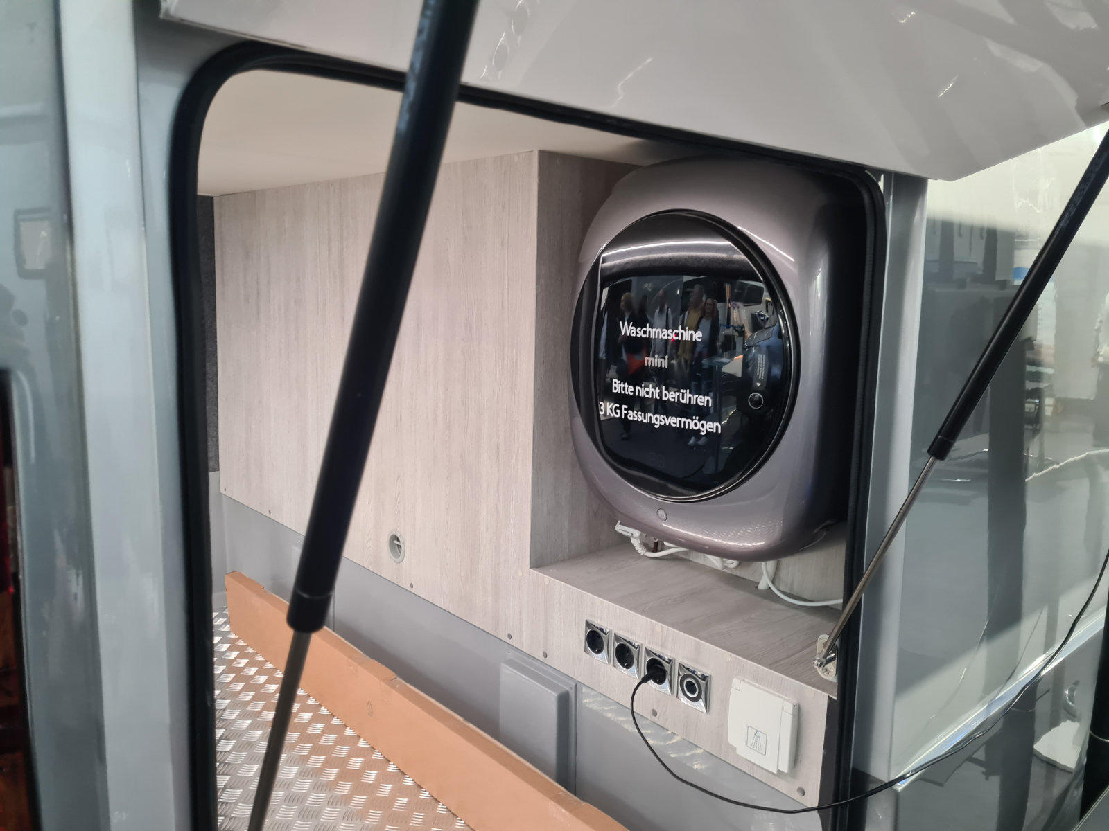 Rundes, wandmontiertes Ger&auml;t mit schwarzer gl&auml;nzender Front und wei&szlig;er Aufschrift; darunter drei Steckdosen in einer wei&szlig;en Leiste; riffeliger Metallboden; schwarze Kabel; zwei Gasdruckd&auml;mpfer