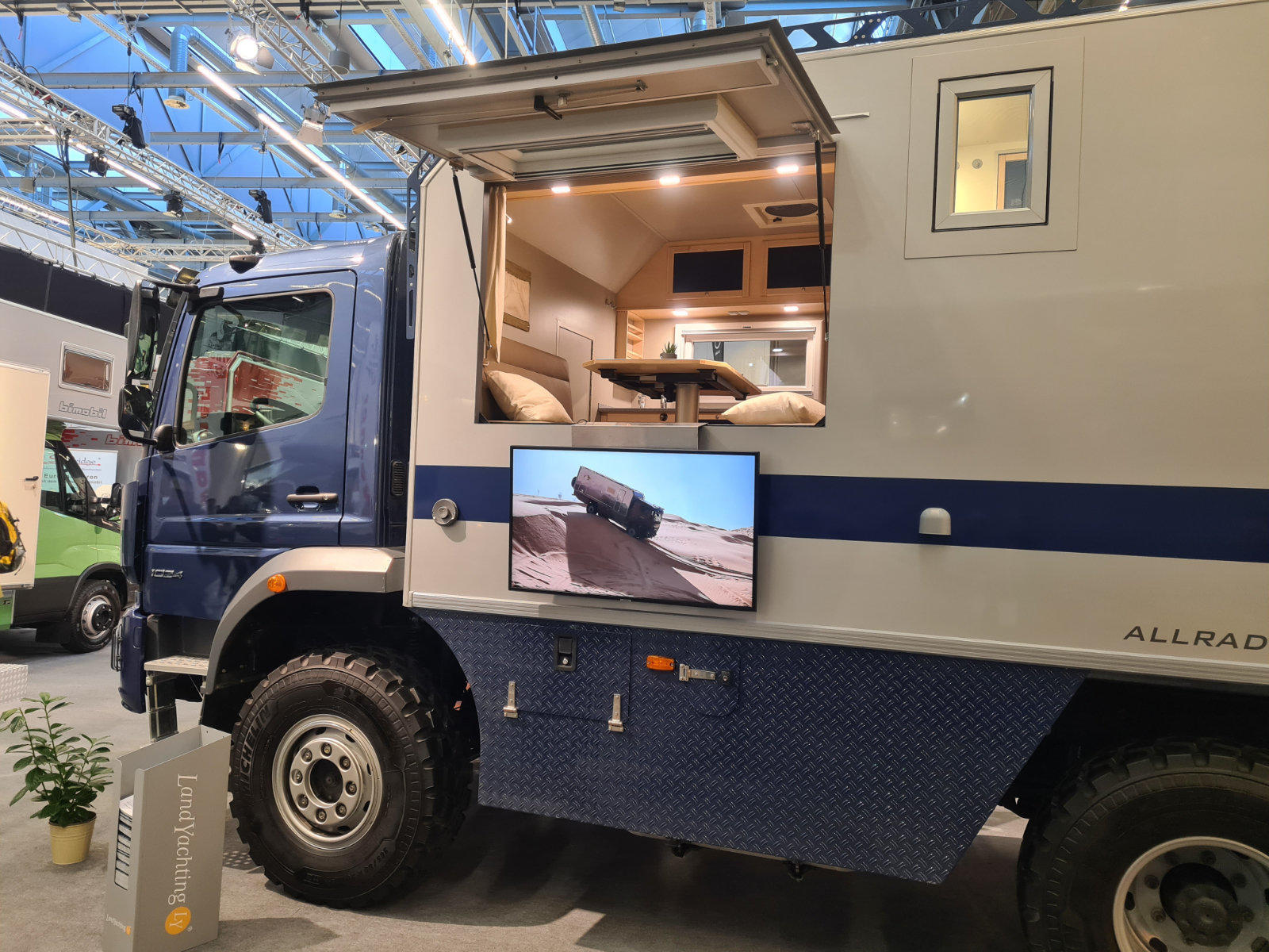 Blauer Lkw mit beigem Wohnmodul in einer &uuml;berdachten Ausstellungsfl&auml;che; seitliche Klappe ge&ouml;ffnet, beleuchteter Innenraum mit Sitzbank, Tisch, Oberf&auml;chern und Kissen sichtbar; au&szlig;en montierter Flachbildschirm unter der Klappe; gro&szlig;es Vorderrad sicht