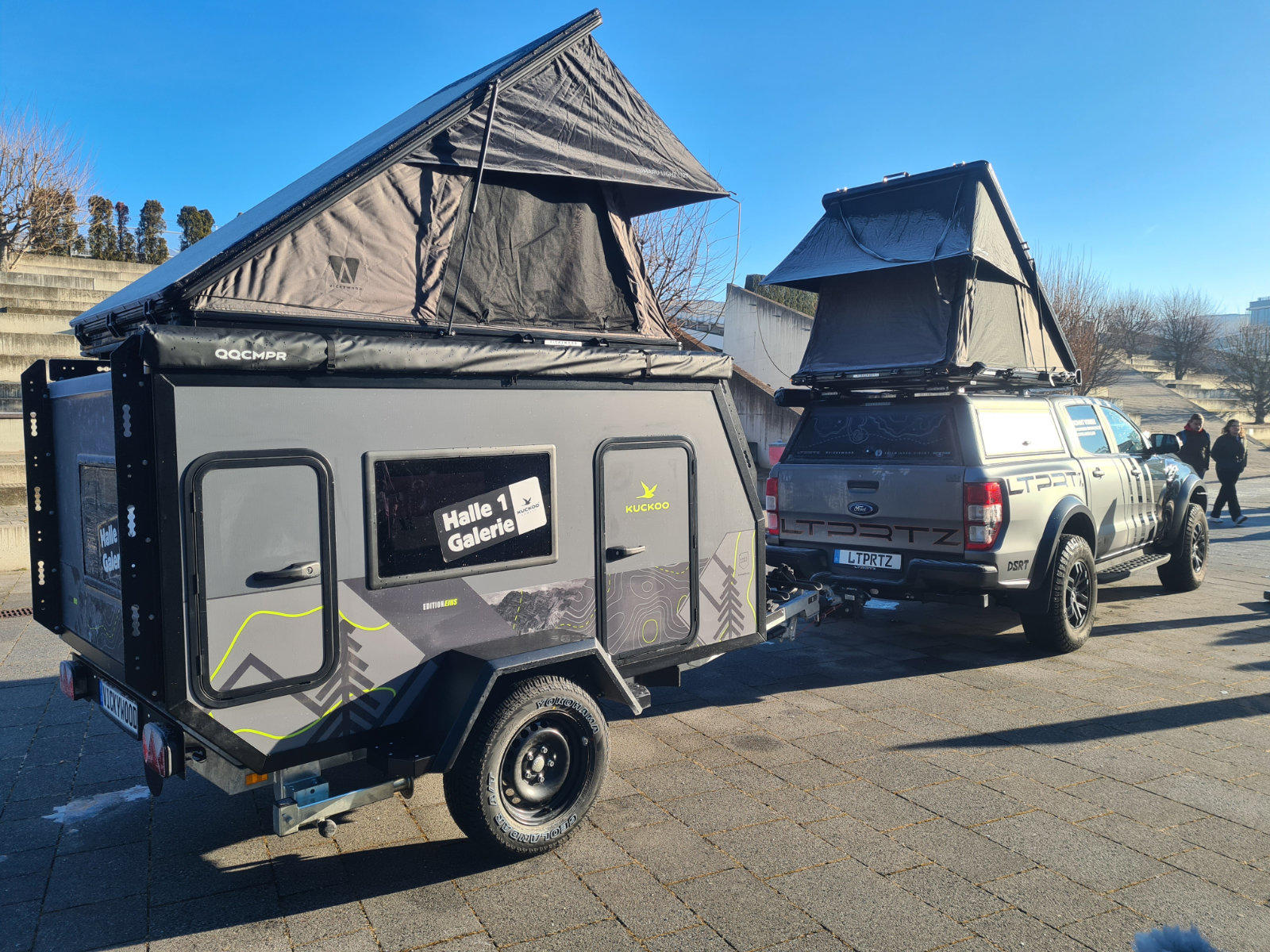Pickup mit aufgeklapptem Dachzelt zieht einen kleinen Anh&auml;nger-Camper mit ebenfalls aufgeklapptem Dachzelt, beide auf gepflastertem Platz unter blauem Himmel