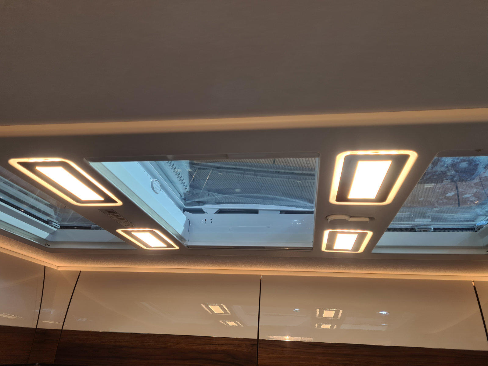 Rechteckiges Fenster in der Decke mit vier rechteckigen Leuchten; darunter gl&auml;nzende wei&szlig;e Schrankt&uuml;ren; durch das Fenster sind Teile eines Fahrzeugs und eines Geb&auml;udes sichtbar