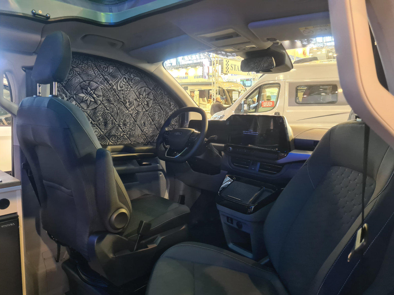 Innenraum eines Fahrzeugs mit zwei Vordersitzen, Lenkrad, Armaturenbrett mit Bildschirm, Mittelkonsole und gemusterter Abdeckung an der Seitenfensterscheibe; durch die Fenster sind weitere Fahrzeuge zu sehen