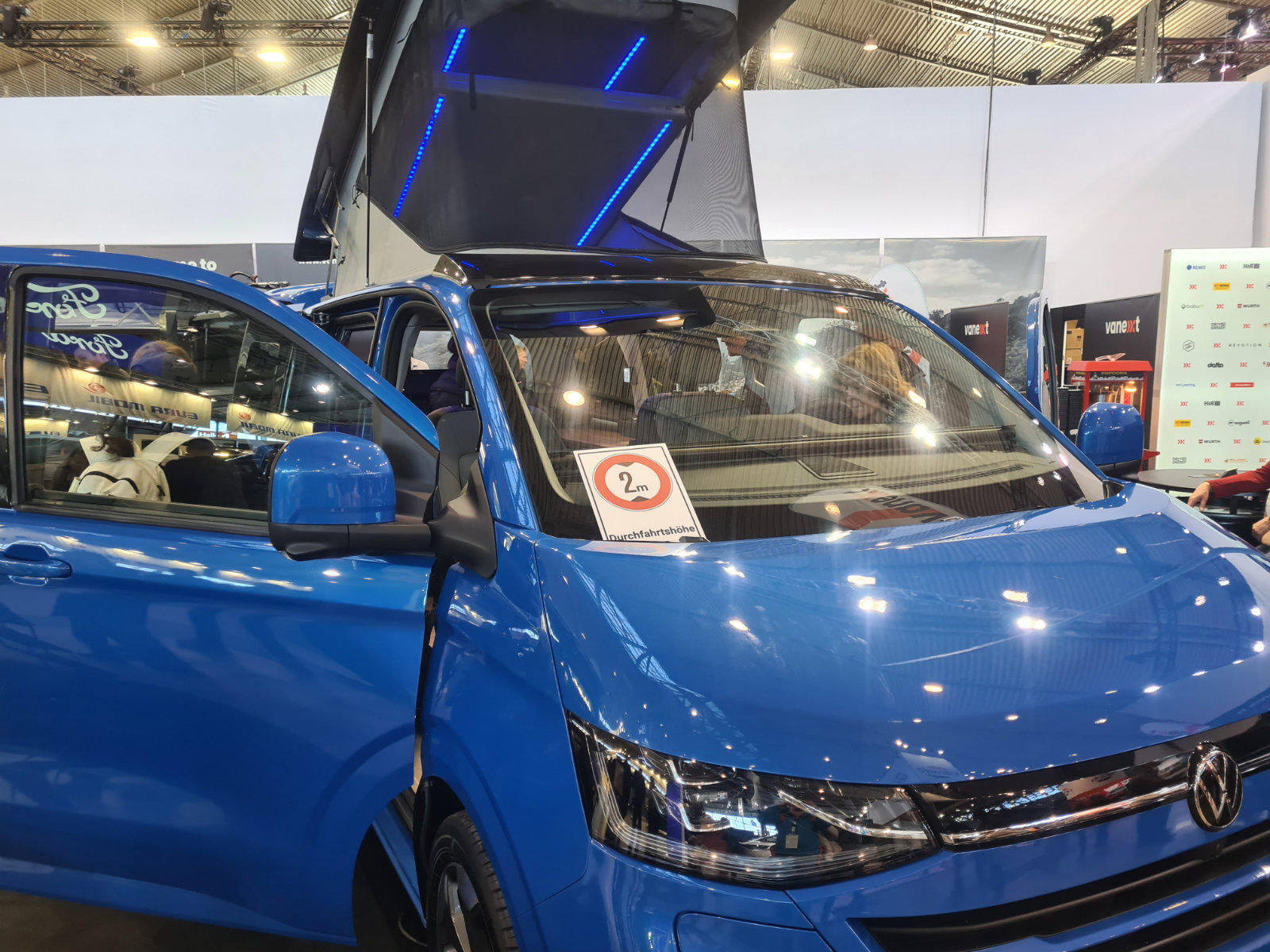 Blauer Volkswagen-Van auf Messe, offene Seitent&uuml;ren, VW-Logo, helle Sitze, runder '2+'-Aufkleber auf Windschutzscheibe