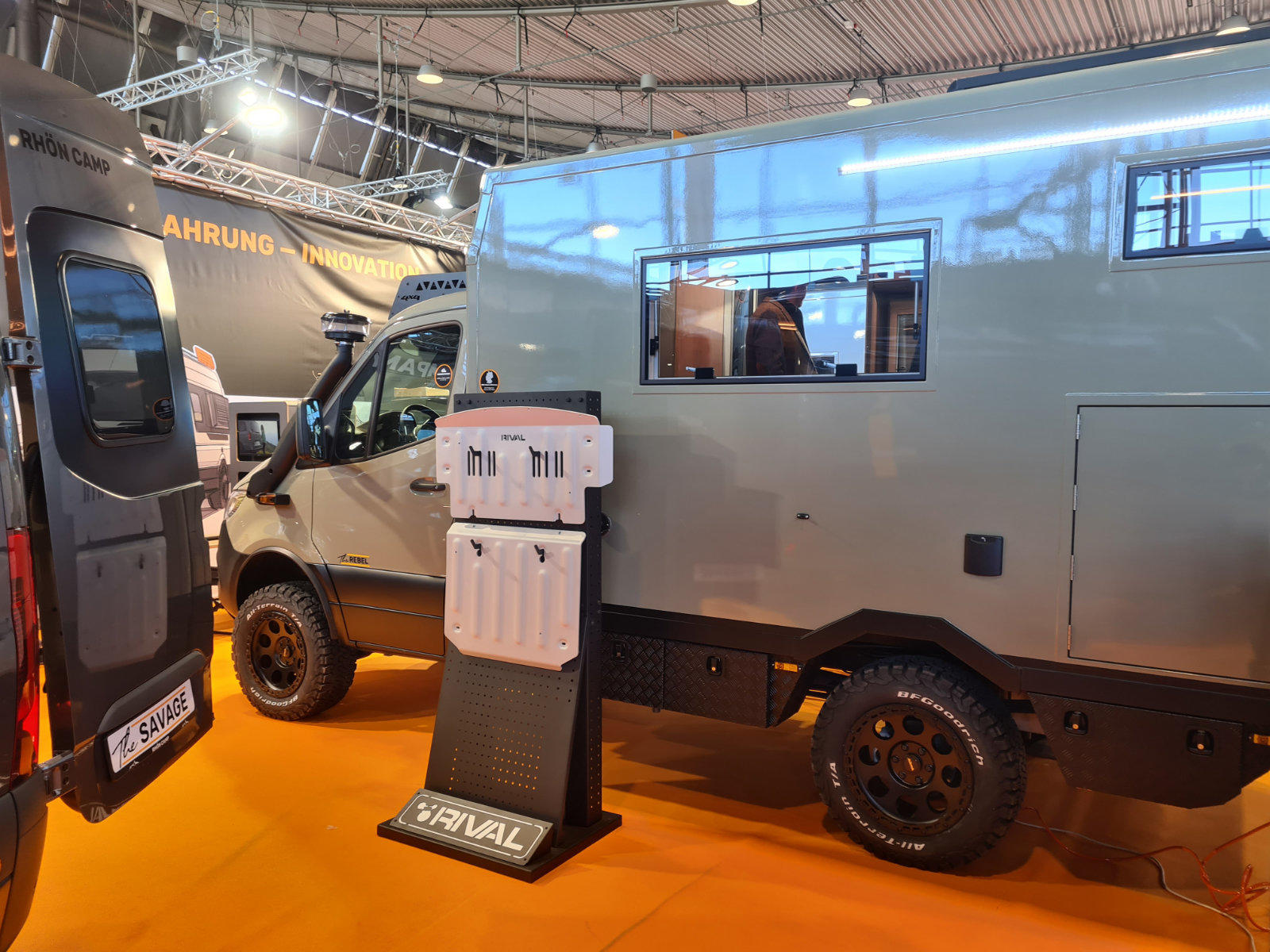 Graues Fahrzeug mit kastenf&ouml;rmigem Aufbau auf orangenem Boden in Ausstellungshalle, ge&ouml;ffnetes Seitenfenster, schwarzer Reifen mit grobem Profil, wei&szlig;er Pr&auml;sentierer auf St&auml;nder vorne, weiteres Fahrzeug links
