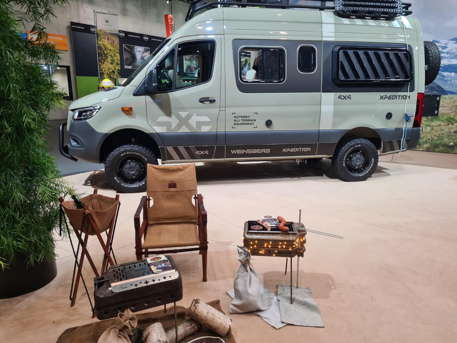 Hellgrauer ausgebauter Van mit Dachtr&auml;ger und Seitenfenstern, schwarze R&auml;der; davor zwei Klappst&uuml;hle, kleiner Campingtisch mit Ausr&uuml;stung, Topfpflanze links