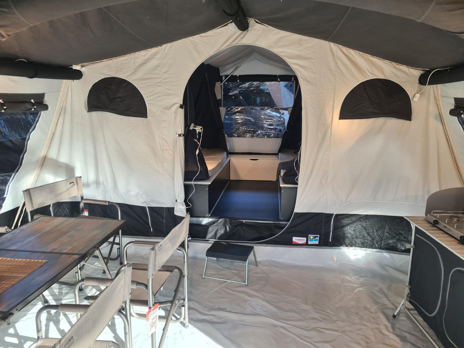 Innenraum eines beigen Zelts mit offenem mittigem Eingang zu einem inneren Abteil, zwei halbkreisf&ouml;rmigen Fenstern, Klapptisch und St&uuml;hlen links, Arbeits- oder Ablagefl&auml;che rechts und kleinem Trittstufenpodest vorn