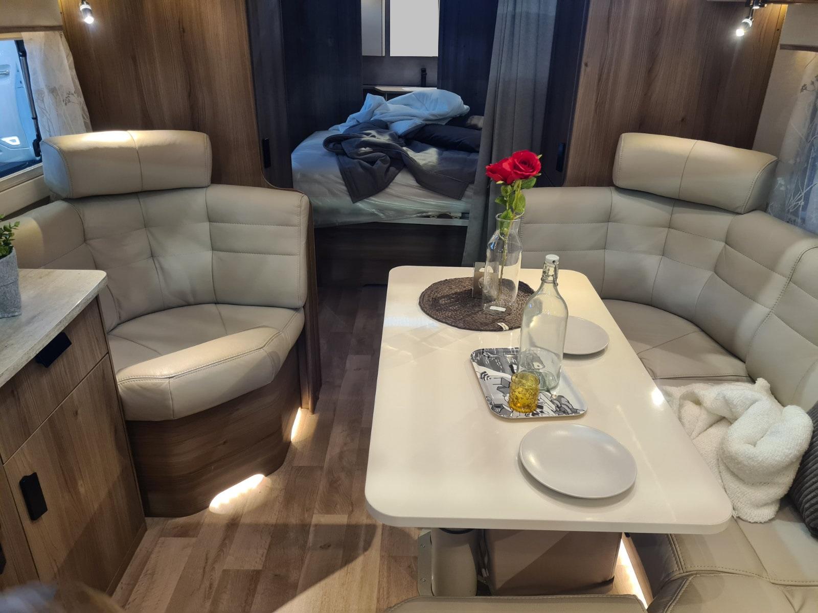 Innenraum mit zwei beigen Polstersitzen, einem wei&szlig;en Tisch mit zwei Tellern, Karaffe, Glas, Tablett und Vase mit roten Blumen; im Hintergrund ein Bett mit aufgeschlagener Bettdecke, Holzvert&auml;felung und Holzboden