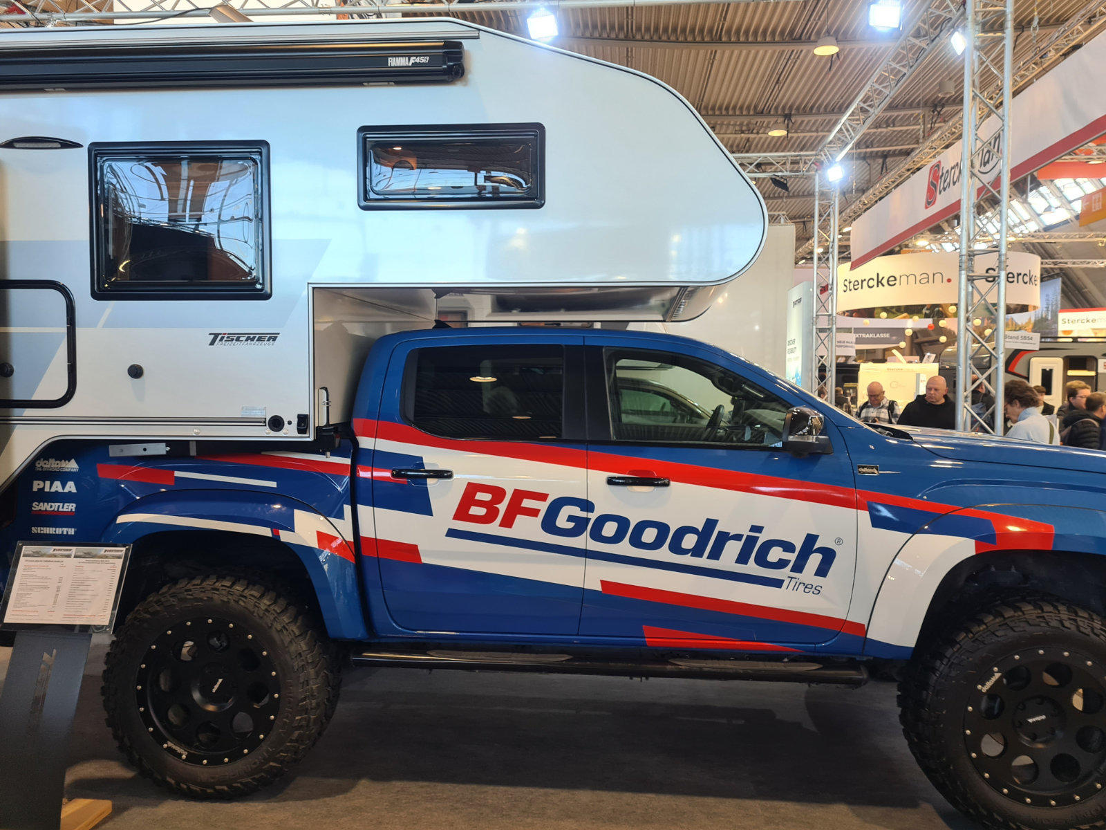 Blauer Pickup mit gro&szlig;em BFGoodrich-Schriftzug an der Seite, wei&szlig;er Wohnaufbau auf der Ladefl&auml;che, gro&szlig;e Gel&auml;ndereifen; im Hintergrund Hallendecke mit Tr&auml;gern und Scheinwerfern, St&auml;nde und Personen