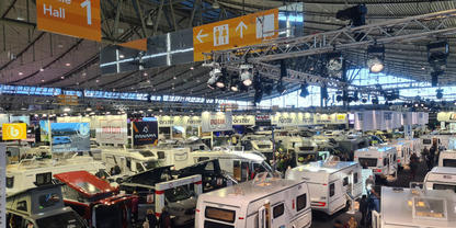 Gro&szlig;e Halle mit vielen Wohnmobilen und Wohnwagen, Personen gehen zwischen den Fahrzeugen, gelbe Wegweiser mit "Halle 1" und Beleuchtung an der Decke
