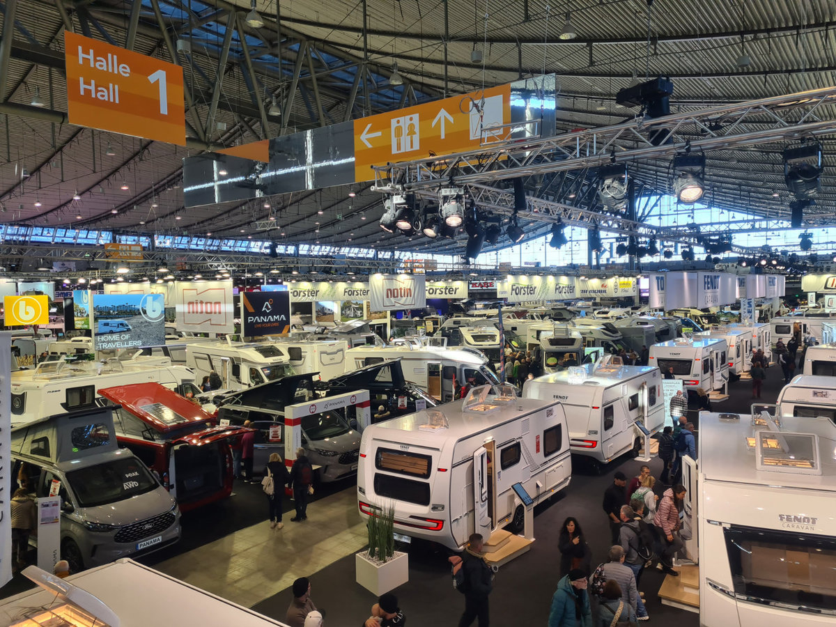 Gro&szlig;e Halle mit vielen Wohnmobilen und Wohnwagen, Personen gehen zwischen den Fahrzeugen, gelbe Wegweiser mit "Halle 1" und Beleuchtung an der Decke