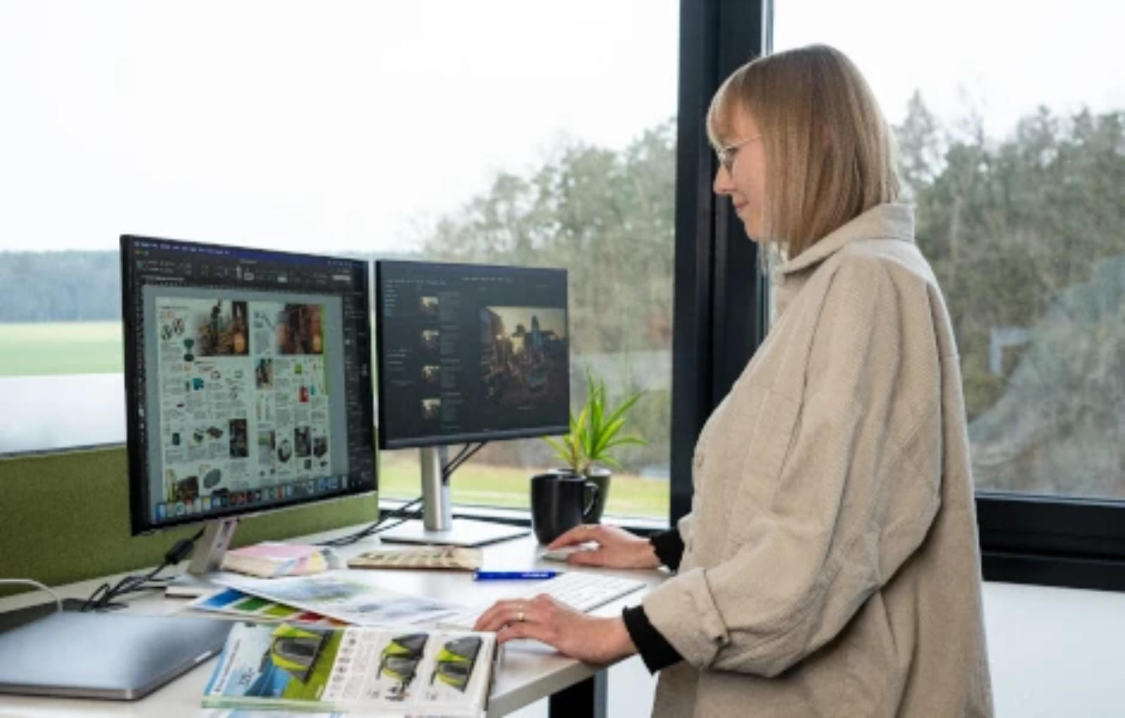 Person steht an Schreibtisch und arbeitet an zwei Monitoren; auf dem Schreibtisch Magazine, Tasse und kleine Pflanze; großes Fenster mit Blick auf Bäume