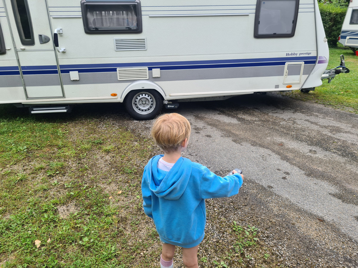 Kind von hinten mit hellblonden Haaren und blauem Kapuzenpullover steht vor einem wei&szlig;en Wohnwagen auf einem grasig-graveligen Platz