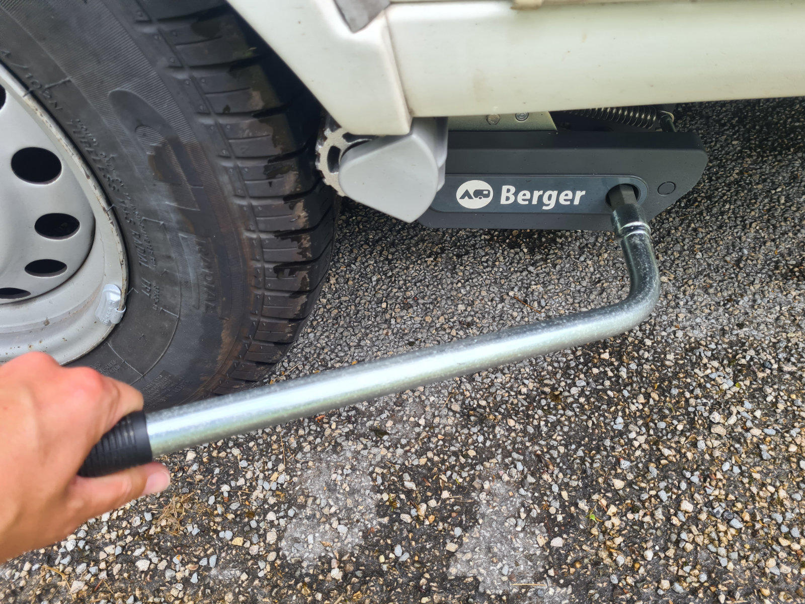 Hand h&auml;lt Metallkurbel am Wagenheber mit Schriftzug "Berger" neben einem Autoreifen, wei&szlig;es Fahrzeug, kiesiger Asphaltboden