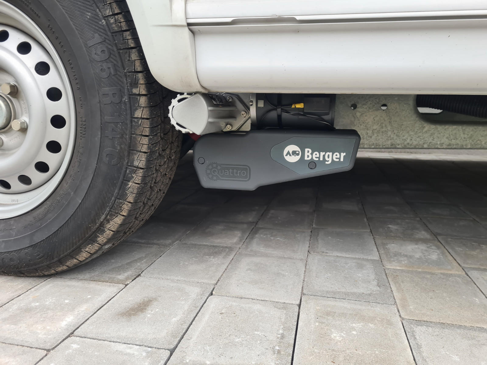 Unterseite eines wei&szlig;en Fahrzeugs; links ein Reifen mit Stahlfelge; darunter ein grauschwarzes Ger&auml;t mit der Aufschrift &bdquo;Berger&ldquo;; graue Pflastersteine
