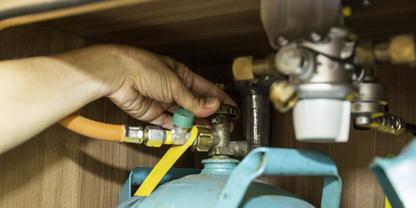 Hand verbindet Schlauch mit Ventil einer blauen Gasflasche in einem Holzschrank