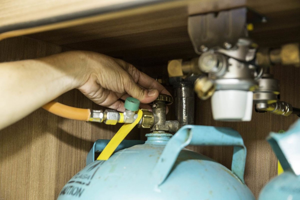Hand verbindet Schlauch mit Ventil einer blauen Gasflasche in einem Holzschrank
