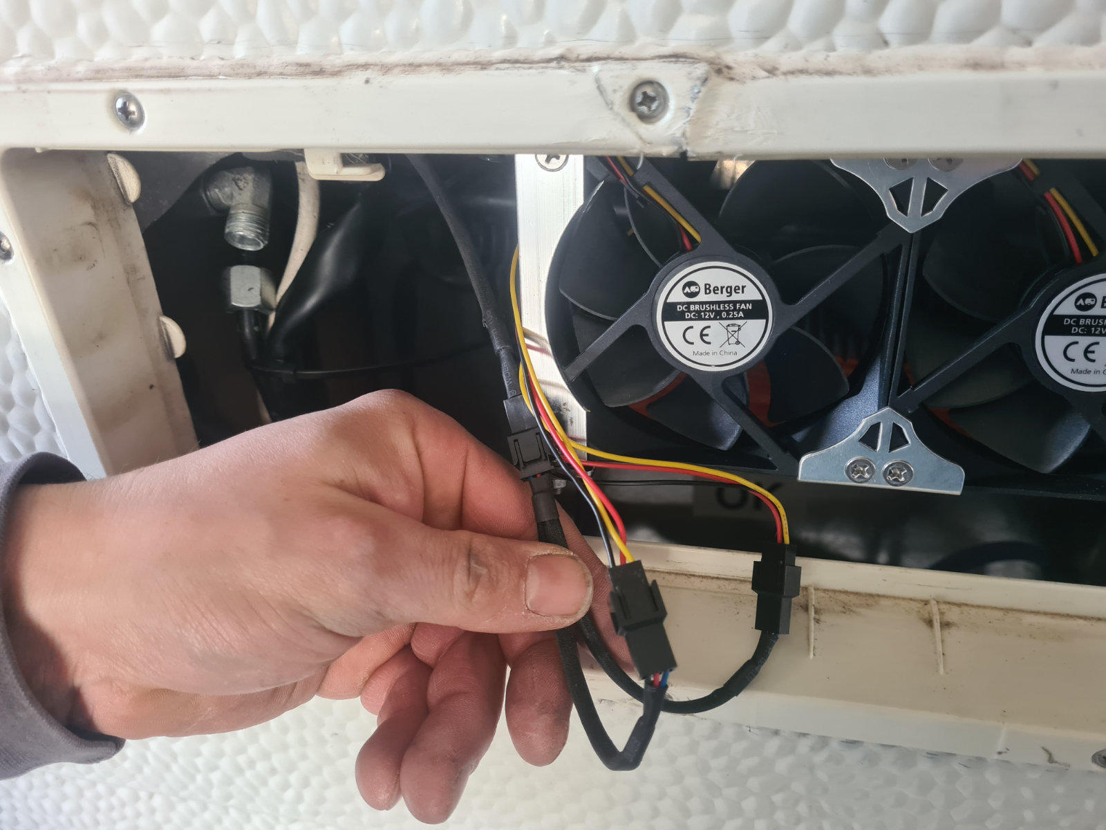 Hand h&auml;lt einen schwarzen Stecker und ein Kabel vor zwei kleinen schwarzen L&uuml;ftern in einem wei&szlig;en Geh&auml;use