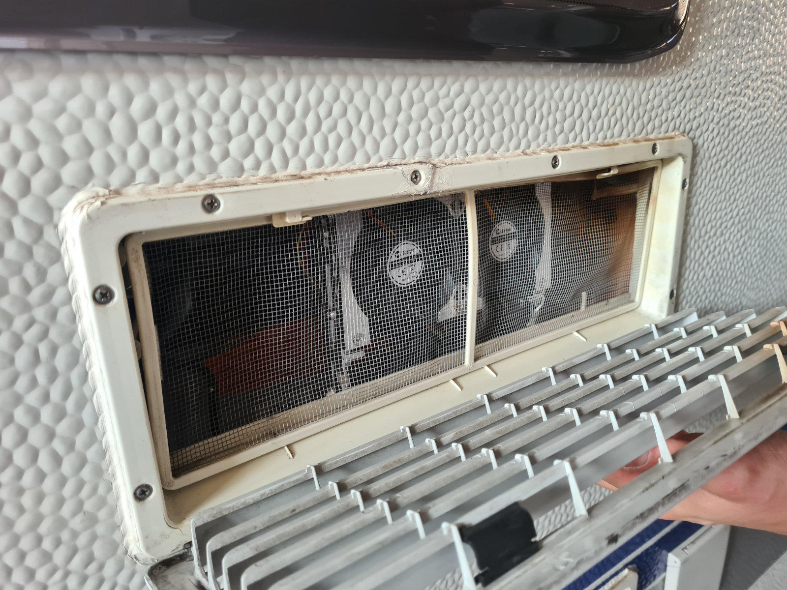 Rechteckige &Ouml;ffnung in wei&szlig;er, strukturierter Oberfl&auml;che mit beigefarbenem Rahmen und Schrauben; hinter schwarzem Drahtgitter zwei runde L&uuml;fter sichtbar; abgenommene graue Kunststoffabdeckung wird von einer Hand gehalten