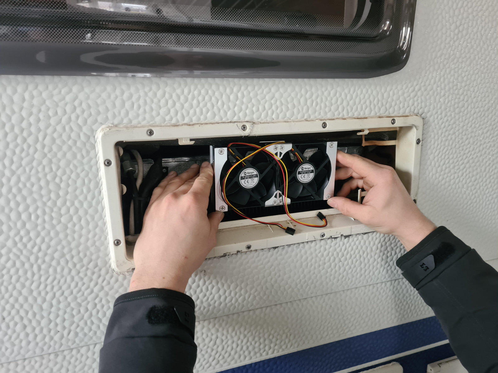 Zwei kleine L&uuml;fter in einem rechteckigen Einbauschacht, zwei H&auml;nde halten die L&uuml;fter, sichtbare Kabel und Schrauben, wei&szlig;e strukturierte Wand