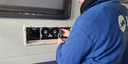 Person in blauer Jacke und beiger M&uuml;tze an der Au&szlig;enwand eines Wohnmobils, h&auml;lt einen Schraubendreher vor einem offenen Wartungsfach mit zwei L&uuml;ftern und sichtbaren Kabeln