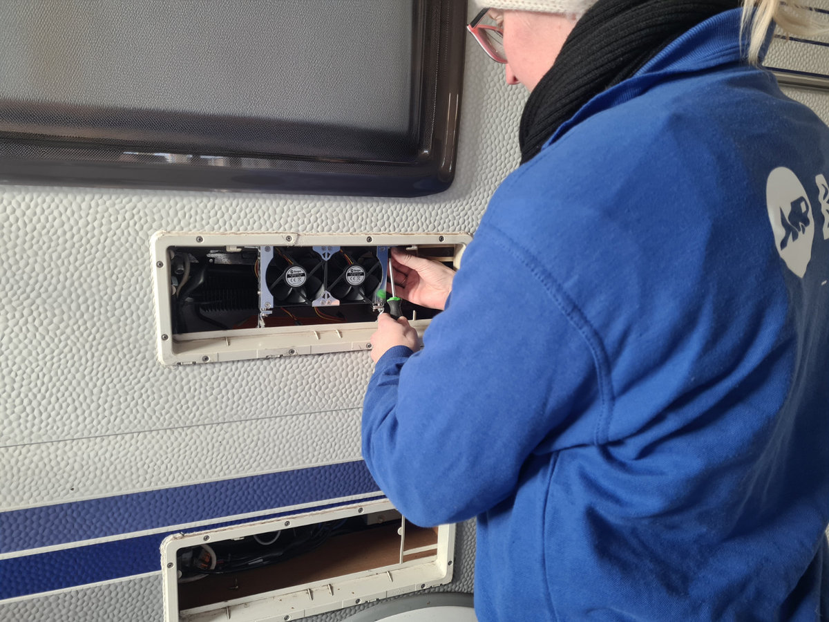 Person in blauer Jacke und beiger M&uuml;tze an der Au&szlig;enwand eines Wohnmobils, h&auml;lt einen Schraubendreher vor einem offenen Wartungsfach mit zwei L&uuml;ftern und sichtbaren Kabeln