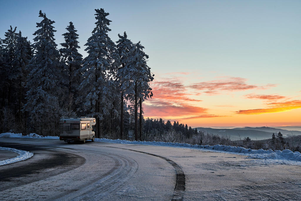 Wohnmobil am verschneiten Stra&szlig;enrand neben einer kurvigen, schneebedeckten Stra&szlig;e; links verschneite Nadelb&auml;ume, rechts entfernte H&uuml;gel und ein farbiger Himmel