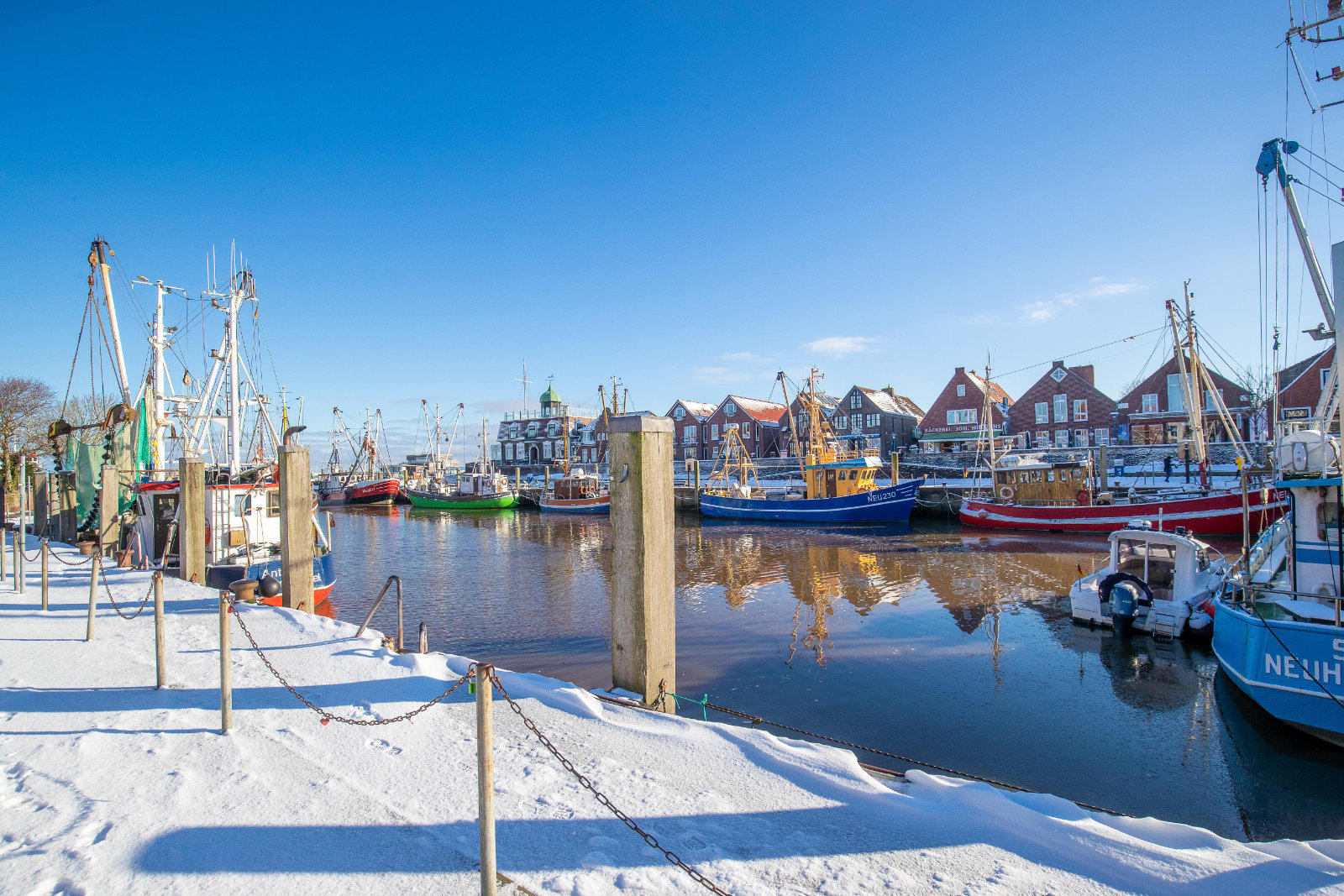 Schneebedeckter Kai mit Pf&auml;hlen, mehrere bunte Fischerboote am Ufer, spiegelndes Hafenwasser und Giebelh&auml;user unter blauem Himmel