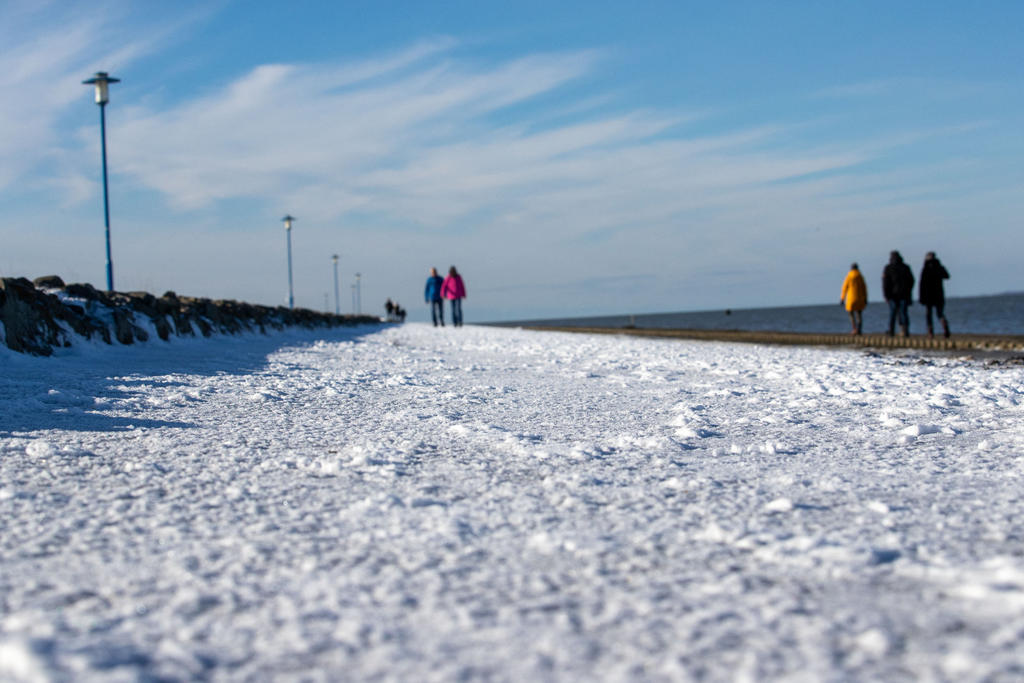 Schneebedeckte Promenade am Meer, Laternen und mehrere Personen unter blauem Himmel