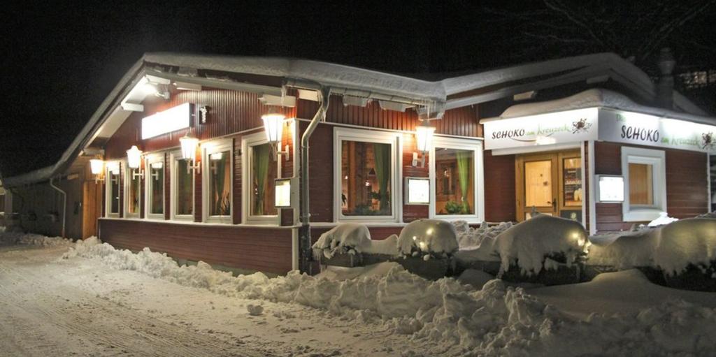 Eingeschossiges rotes Holzgeb&auml;ude mit wei&szlig;en Zierleisten, beleuchteten Fenstern und beleuchteter Beschilderung, schneebedeckte B&uuml;sche und Boden, Nachtaufnahme