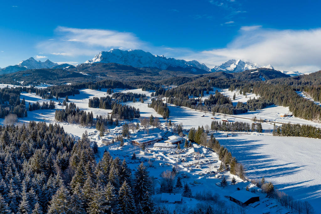 Verschneites Dorf in einem Tal mit Nadelwald, schneebedeckten Feldern, schneebedeckten Bergen und blauem Himmel mit Wolken