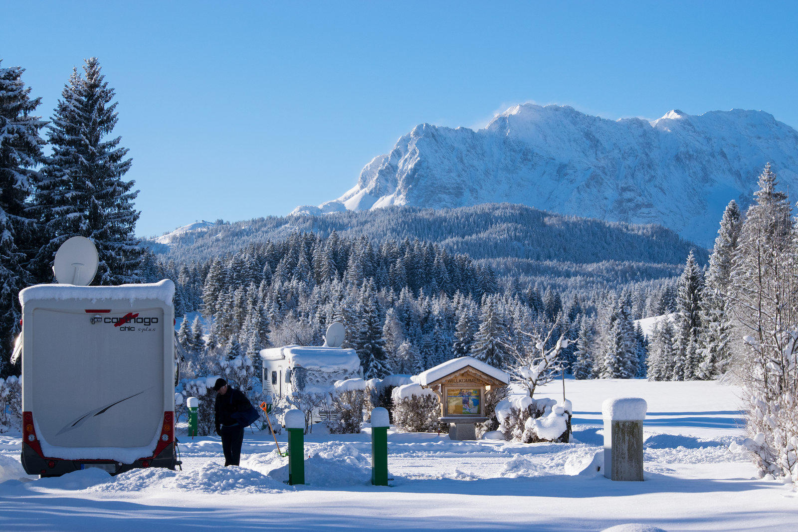 Schneebedeckte Landschaft mit abgestelltem Wohnmobil links, einer gehenden Person daneben, kleinem verschneitem Holzhaus in der Bildmitte, schneebedeckte Tannen, schneebedeckter Berg im Hintergrund und blauer Himmel