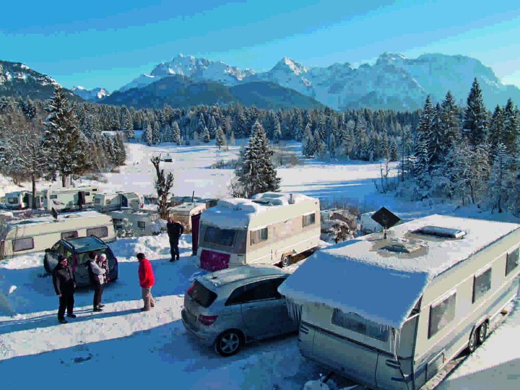 Mehrere schneebedeckte Wohnmobile und Autos auf offenem Platz, einige Personen, Tannenb&auml;ume und verschneite Berge im Hintergrund, blauer Himmel