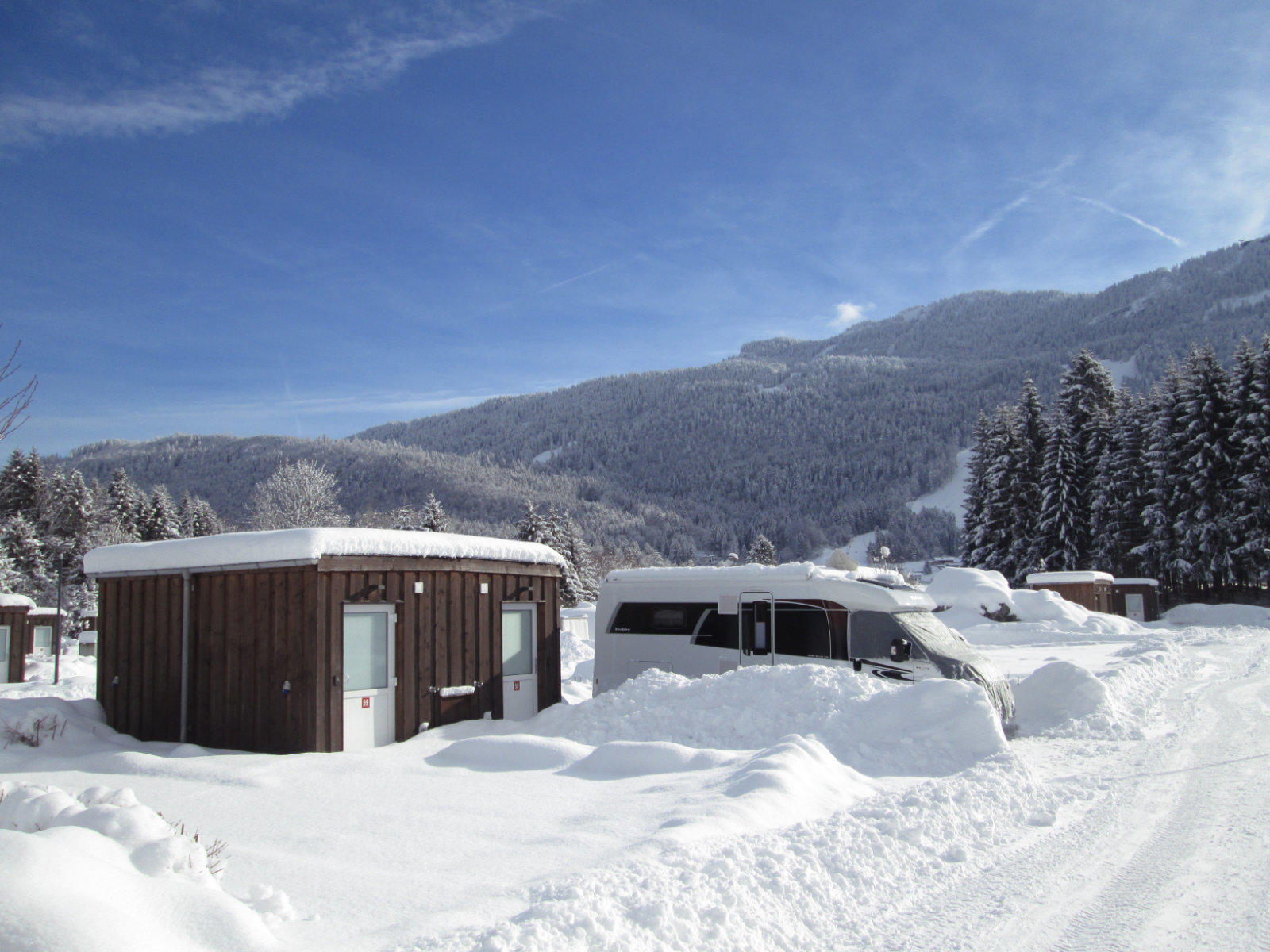 Schneebedeckte Holzh&uuml;tten und ein Wohnmobil auf einem verschneiten Platz, dahinter Tannen und schneebedeckte Berge unter blauem Himmel