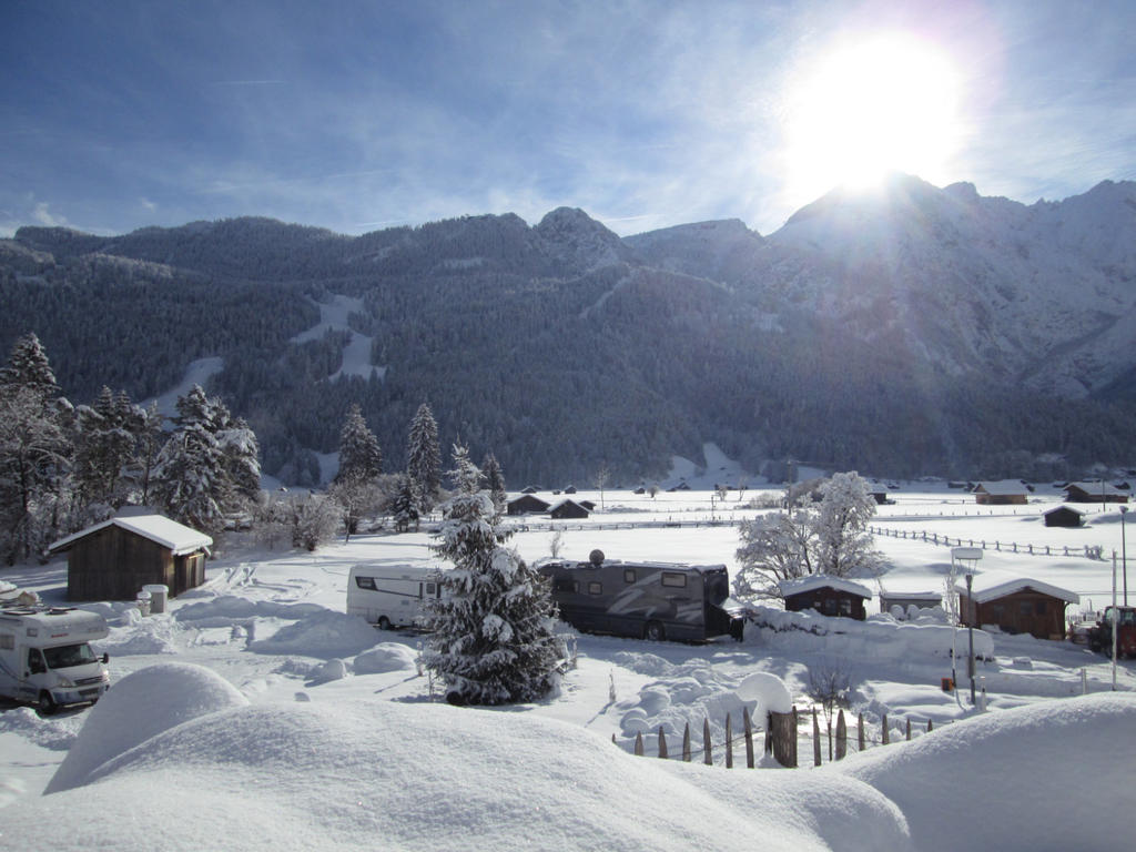 Schneebedeckte H&auml;user, Fahrzeuge, B&auml;ume und Z&auml;une in einem verschneiten Tal, im Hintergrund sonnenbeschienene Berge und blauer Himmel