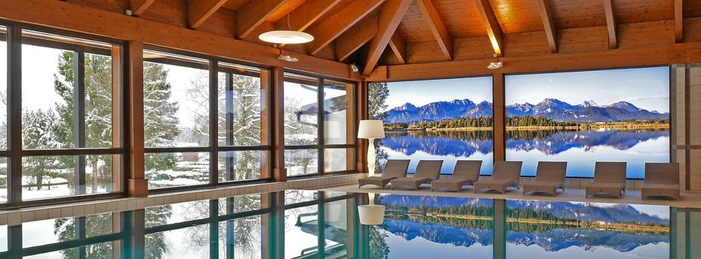 Innenpool mit sichtbarer Holzdecke und Balken, Liegen am Beckenrand, gro&szlig;e Fenster mit Blick auf schneebedeckte B&auml;ume und Berge, Spiegelung im Wasser