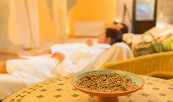 Schale mit getrockneten Kr&auml;utern auf einem gemusterten Tisch im Vordergrund; im Hintergrund unscharf eine Person im wei&szlig;en Bademantel auf einer Liege in einem warm beleuchteten Raum, Kissen und Pflanzen sichtbar