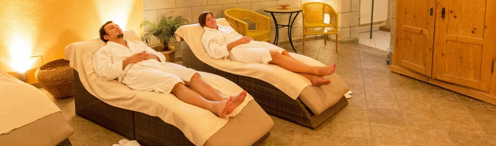 Zwei Personen in wei&szlig;en Badem&auml;nteln liegen auf gepolsterten Liegen in einem Raum mit warmem Licht, Holzm&ouml;beln, kleinem Tisch mit zwei St&uuml;hlen, Pflanze und gefliestem Boden