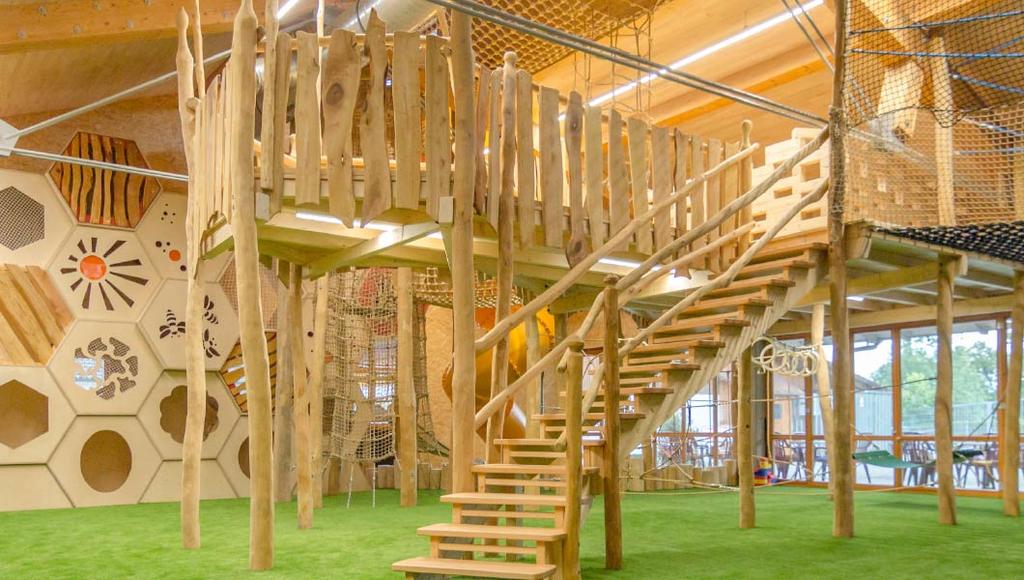 Innenraum mit gro&szlig;er Holzkonstruktion: Treppe zu erh&ouml;hter Plattform, h&auml;ngende Holzlatten, Netze am Gel&auml;nder, gr&uuml;ner Teppichboden, sechseckige Wandnischen, gro&szlig;e Fenster, Tische und St&uuml;hle