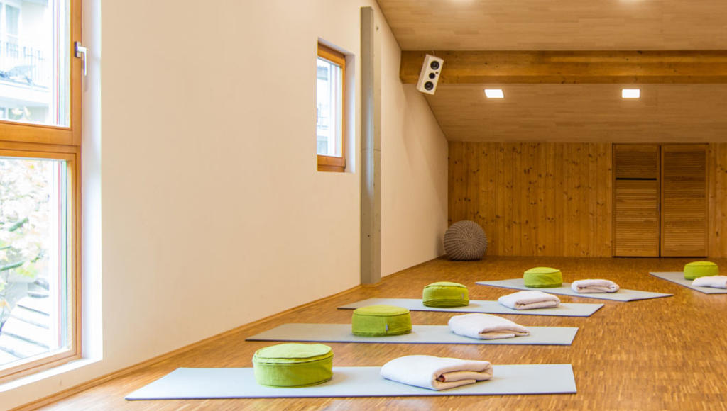 Heller Raum mit Holzfu&szlig;boden und Holzwand, Fenster, mehrere blaue Matten, gr&uuml;ne runde Polster, gefaltete Handt&uuml;cher und ein gro&szlig;er grauer Ball in der Ecke