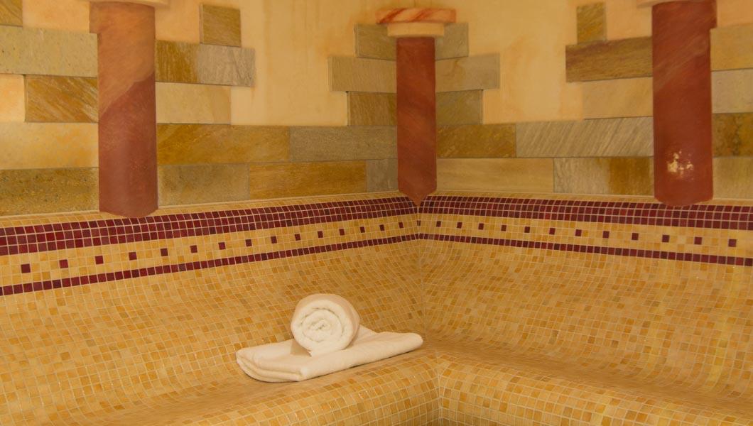 Mosaikverkleidete Sitzfl&auml;che mit aufgerolltem wei&szlig;em Handtuch; dahinter gestreifte Wandfliesen und zwei runde S&auml;ulen